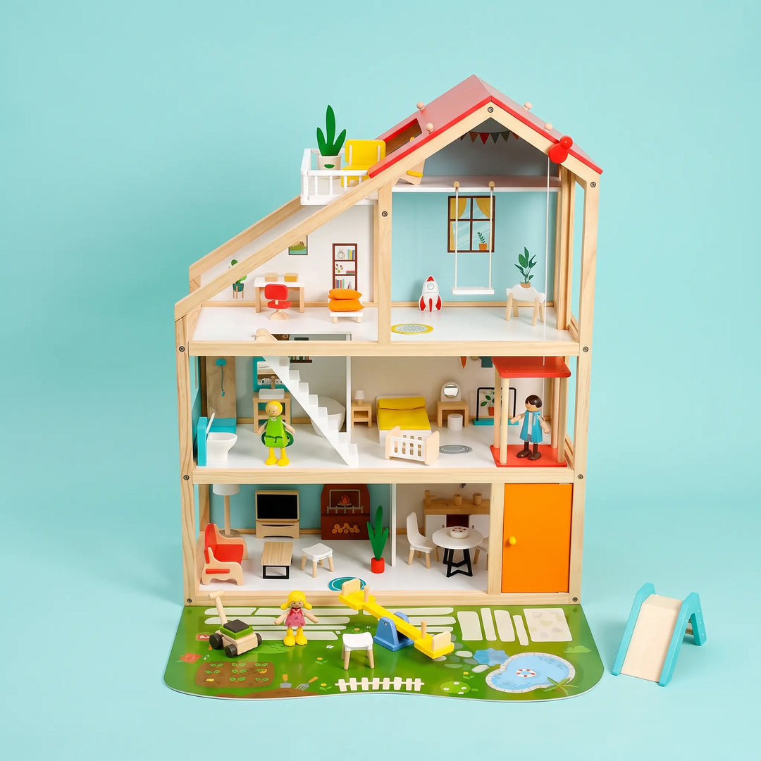 Three-story wooden dollhouse with a red pitched roof, featuring detailed interior rooms, furniture, and posable figures, set on a garden mat with a slide and pond.