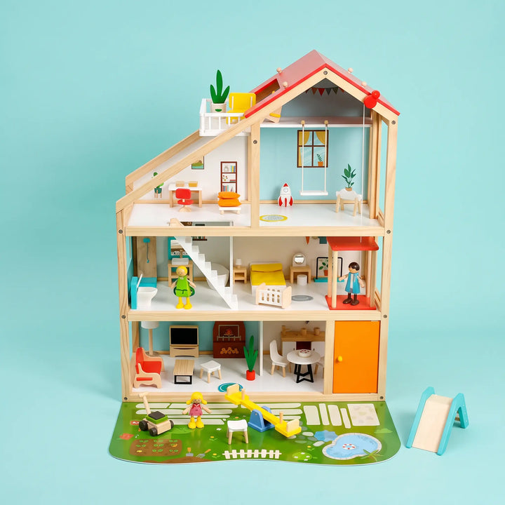 Three-story wooden dollhouse with a red pitched roof, featuring detailed interior rooms, furniture, and posable figures, set on a garden mat with a slide and pond.