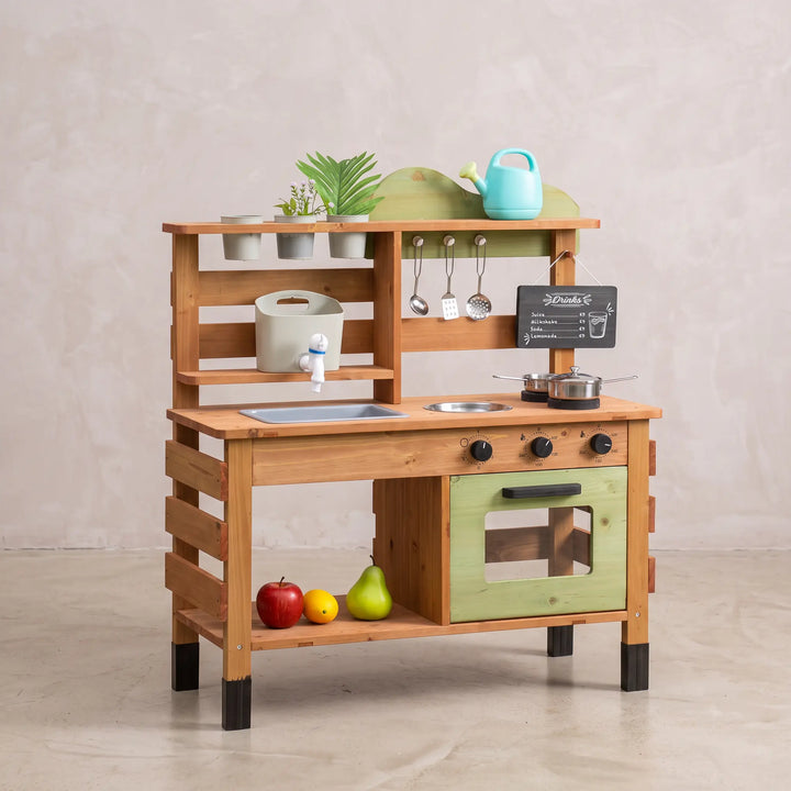 Wooden outdoor kitchen playset with sink, stovetop, oven, and chalkboard, set in a garden with plants and accessories. Features a light green oven door, blue watering can, and play fruit.