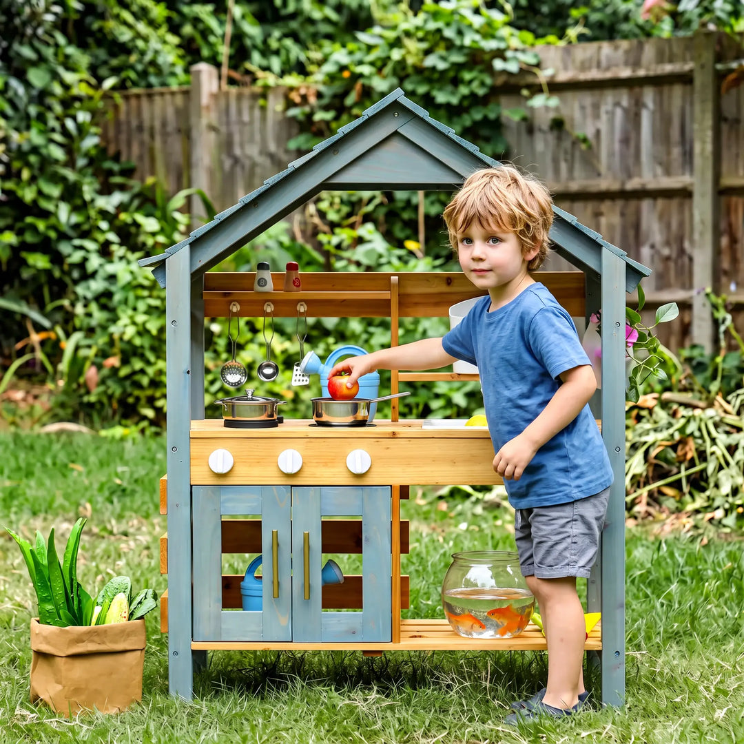 A young boy plays with a charming wooden outdoor kitchen set on a sunny day, featuring a light blue roof and doors, natural wood details, and various cooking accessories.