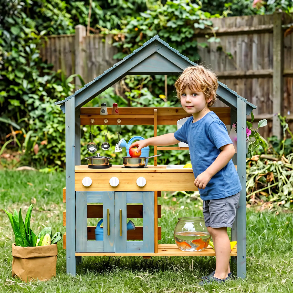 A young boy plays with a charming wooden outdoor kitchen set on a sunny day, featuring a light blue roof and doors, natural wood details, and various cooking accessories.