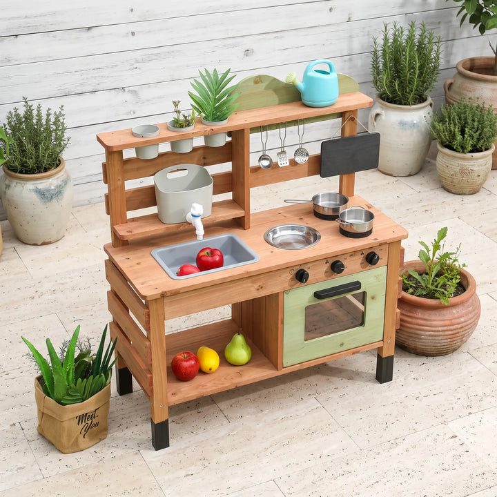 A wooden outdoor kitchen playset with a light green oven door and clear window, set in a garden with plants and accessories like a blue watering can and chalkboard menu.