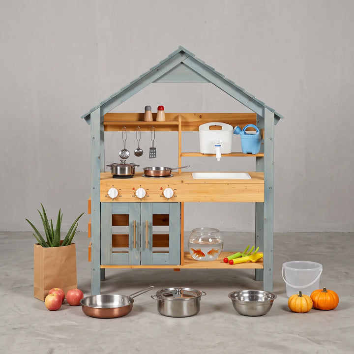 Wooden outdoor play kitchen with a light blue slatted roof and blue cabinet doors, featuring natural wood shelves, a white sink with a blue-ringed faucet, and four burner knobs. Includes play accessories like pots, pans, and utensils, displayed with a plant, apples, and pumpkins.